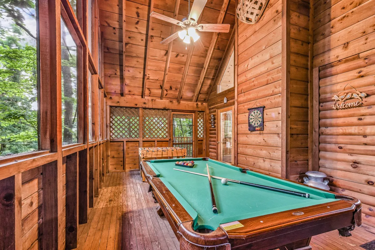 A welcoming entertainment space featuring a pool table, wooden walls, a dartboard, and a ceiling fan. The cozy ambiance is accented by a 'welcome' sign, suggesting a relaxed atmosphere ideal for games and socializing.