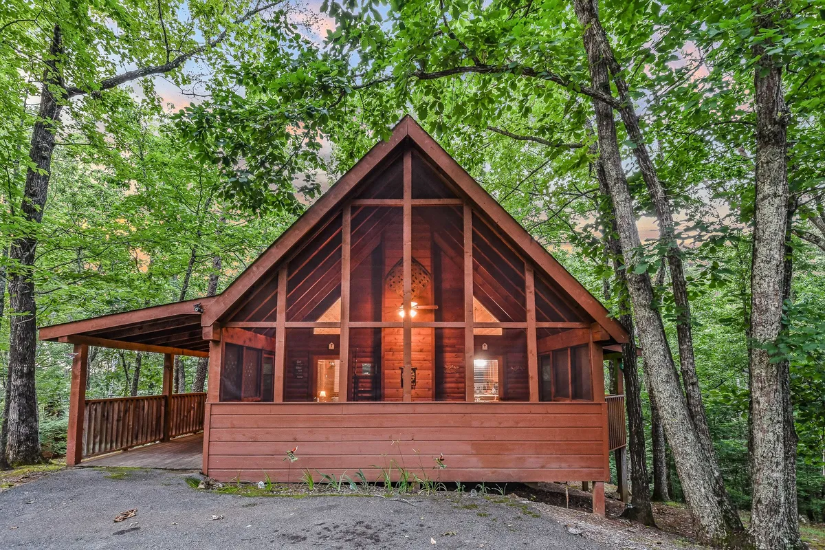 This image captures the A-frame structure of the cabin, large windows that offer views of the surrounding trees, and a spacious wooden deck. The natural setting is serene, showcasing the cabin as a base for both relaxation and adventure.