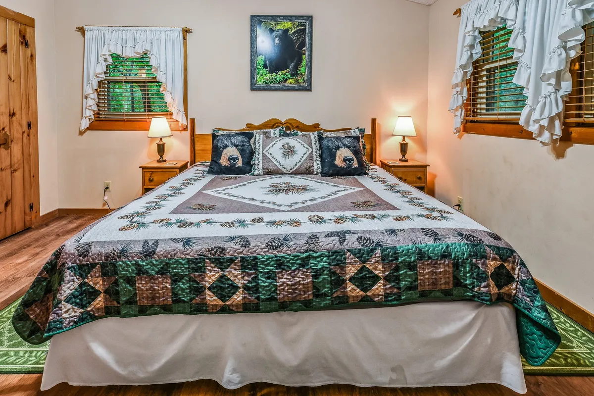 A serene king bedroom with a bed adorned with a quilt and bear-themed pillows, complemented by tasteful wall art and softly lit nightstands. The overall atmosphere is tranquil and restful, echoing the promise of peaceful nights and cozy relaxation.