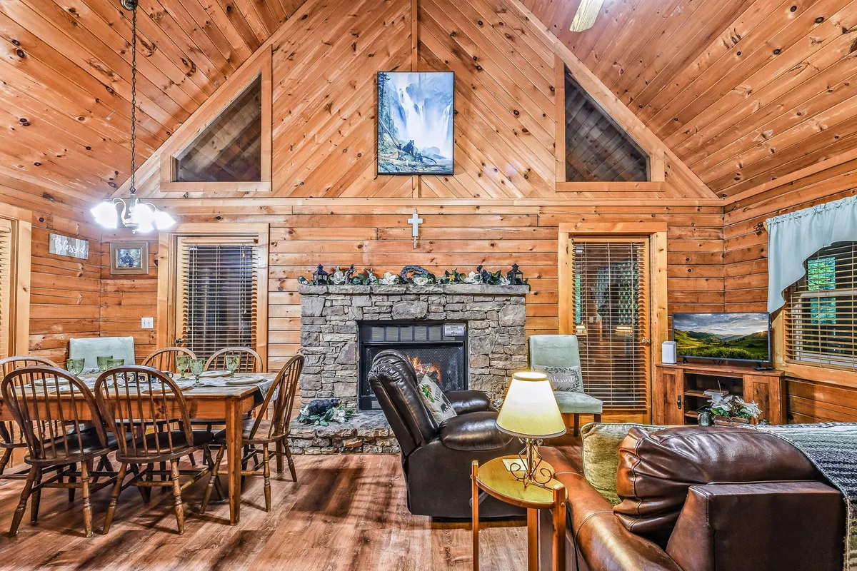 An open-plan room showcasing a stone fireplace, spacious dining table, wooden beams, large windows, and artwork. The room is bathed in warm light, with cozy furnishings inviting guests to relax, dine, or enjoy a game night.