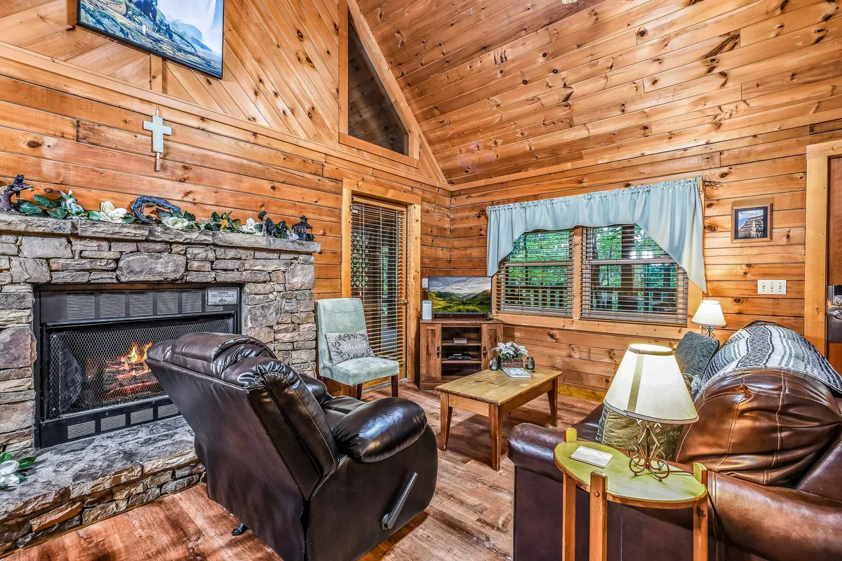 A cozy living room featuring a majestic stone fireplace, wooden walls, comfortable seating, a decorative cross, and large windows letting in natural light. The inviting arrangement is perfect for family gatherings, quiet reading moments by the fire, or enjoying the view of the outdoors.