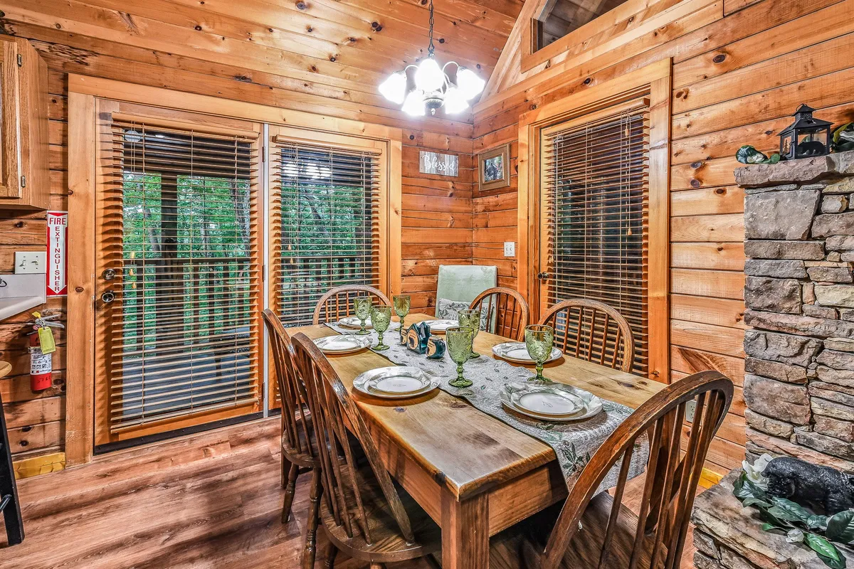 This inviting dining area features a six-person wooden dining table, a cozy fireplace, large windows bringing in natural light, and wooden walls that enhance the cabin’s rustic charm. The table is set for a gathering, creating a setting perfect for shared meals and celebrations.