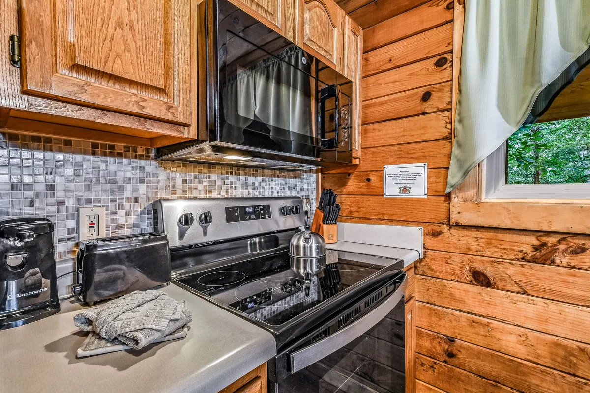 A close-up look at the kitchen’s cooking area, featuring a stainless steel stove, wooden cabinetry, a colorful mosaic backsplash, a kettle, and a window that offers views of greenery outside. The space is bathed in natural light, emphasizing its inviting and functional qualities.