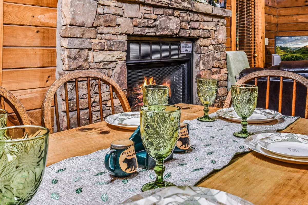 A rustic-chic dining area with a wooden table set for a meal, green glassware, a decorative plate display, and a visible stone fireplace in the background. The setup radiates a sense of togetherness and is ideal for both family dinners and intimate gatherings.