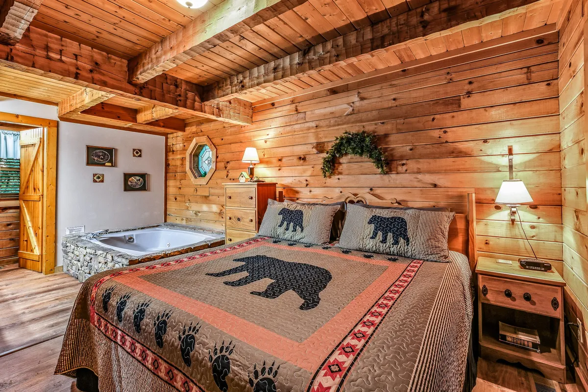A cozy interior bedroom with a bear-themed quilt on the bed, wooden walls and ceiling, decorative wall art, and a jacuzzi tub in the corner. The space radiates warmth and comfort, inviting guests to unwind after a day outdoors.