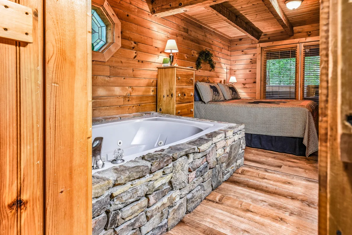A romantic and tranquil suite featuring a wooden bed, a luxurious jacuzzi tub, and a rustic stone wall. The room offers a cozy sanctuary with views through the window, inviting relaxation and indulgent soaks in the spa-inspired bath.