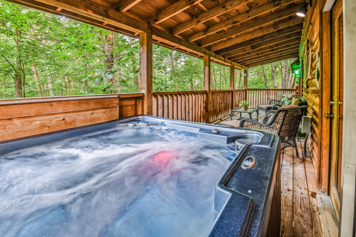 A tranquil exterior shot featuring a private hot tub on a wooden deck surrounded by lush greenery. The setting includes comfortable seating, providing a peaceful and secluded atmosphere that invites couples to relax together under the open sky.