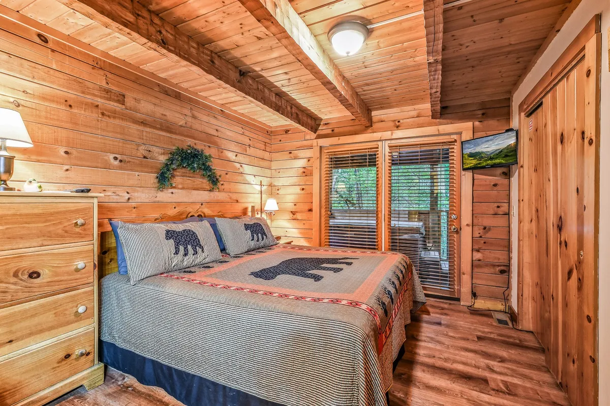 A rustic bedroom featuring a bed with bear-themed bedding, a wooden dresser, and large windows with blinds. The windows allow for peaceful views of the surrounding woods, blending the indoor space with nature just outside.